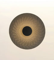 A circular woven basket or mat with a textured geometric pattern, featuring a dark brown center and a light brown outer rim, set against a plain off-white background.