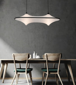 A modern pendant light with a white curved shade and black frame hangs above a wooden dining table with two chairs in a minimalist room with a textured gray wall.