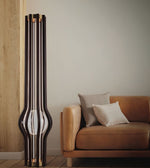 A tall, sculptural black floor lamp with multiple vertical glass panels and a rounded base, standing next to a tan leather sofa with two neutral throw pillows in a minimalist living room setting.