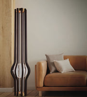 A tall, sculptural black floor lamp with multiple vertical glass panels and a rounded base, standing next to a tan leather sofa with two neutral throw pillows in a minimalist living room setting.