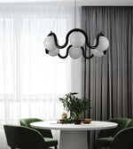 A modern black wavy pendant light with five white spherical glass shades hangs over a round white dining table with green chairs in a contemporary room with sheer white curtains and dark gray drapes.