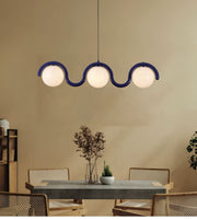 A modern three-light wavy pendant chandelier with frosted white globe shades and a dark blue frame, hanging above a dining table in a warm beige room.