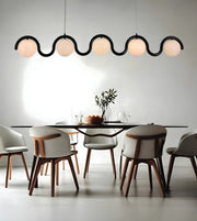 A modern dining room scene with a long black wavy chandelier featuring six glowing frosted globe shades hanging above a dark rectangular table and six upholstered chairs with light gray fabric and medium brown wooden legs.
