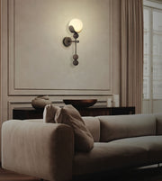 A modern wall-mounted sconce with a frosted round globe and dark metal accents, mounted on a beige paneled wall above a beige sofa in a styled living room.