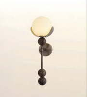 A modern wall sconce light fixture with a glowing frosted glass globe, dark bronze metal accents, and a circular wall mount, shown against a soft light beige background.