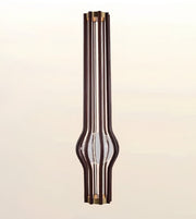 A modern vertical pendant light fixture with multiple dark brown wood-grain rods, brass-toned metal caps, and a clear glass center tube, shown against a soft beige gradient background.