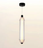 A modern cylindrical pendant light with a ribbed clear glass shade, black ceiling canopy and cord, shown against a soft off-white background.