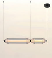 A modern two-light linear pendant lamp with clear cylindrical glass shades and warm white LED tubes, suspended by thin black cords from a black ceiling canopy against a plain off-white background.