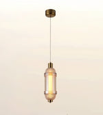 A modern cylindrical pendant light with a textured clear glass shade and brushed brass metal accents, suspended from a matching ceiling canopy against a soft warm gradient background.