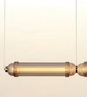 A modern cylindrical pendant light fixture with a brushed gold metal frame, textured end caps, and a ribbed clear glass shade, suspended against a plain off-white background.