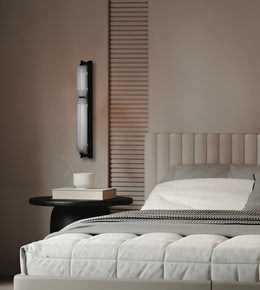 A modern bedroom scene featuring a black wall-mounted sconce with two vertical frosted glass shades, styled on a beige wall next to a bed with white bedding and a black side table.