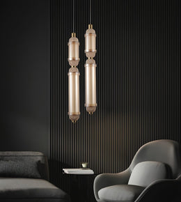 Two modern pendant lights with elongated cylindrical glass shades and brushed brass-toned metal accents, hanging in a dark contemporary room with a ribbed black wall, gray sofa, and curved gray armchair.