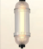 A cylindrical pendant light fixture with a warm white illuminated glass shade, featuring textured clear glass caps and silver-toned metal bands, shown against a plain light background.