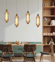 Four vintage-style pendant lights with ribbed glass and brass-toned metal accents hang over a dining table in a modern, sunlit room with wooden chairs and a bookshelf.