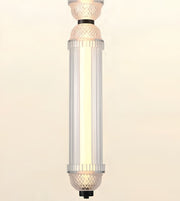 A modern cylindrical pendant light fixture with a ribbed clear glass shade and textured glass caps, glowing with a warm light against a plain beige background.