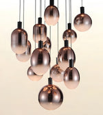 A cluster of modern pendant lights with metallic rose gold glass globes and teardrop shapes hanging from black cords against a warm off-white background.