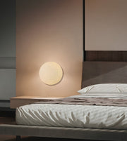 A modern minimalist bedroom featuring a round, flush-mounted warm white wall sconce on a beige wall, next to a low wooden nightstand and a bed with white textured bedding.