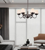 A modern six-light chandelier with a dark metal frame and warm white drum shades hangs in a contemporary living room with a sofa, armchair, and large sheer curtains.