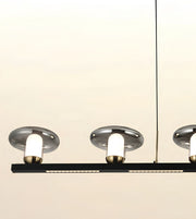 Modern linear pendant light fixture with three illuminated cylindrical bulbs in brushed silver shades and a black horizontal bar, shown against a plain light beige background.