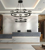 A modern multi-tier circular chandelier with black metal rings and clear glass shades hanging in a contemporary lobby with a marble reception desk and white ceiling.