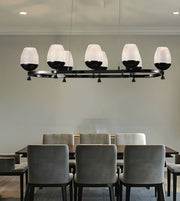 A modern 7-light linear chandelier with textured white glass drum shades and black metal arms, suspended over a dining table with gray chairs in a dining room.