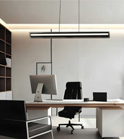 A modern minimalist office setup featuring a long, slim rectangular LED pendant light hanging over a light wood desk with a white computer monitor, black office chair, and laptop.