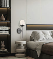 A modern wall-mounted sconce with a white frosted glass shade and a dark metal base, mounted on a bedroom wall next to a bed and a small round side table.