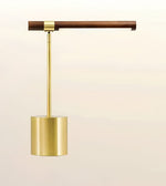 A modern pendant light fixture with a brushed brass cylindrical shade, a slim brass stem, and a dark brown wooden horizontal bar, shown against a soft beige background.
