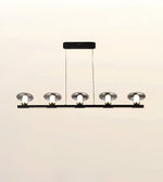 A modern 5-light linear ceiling chandelier with a matte black horizontal bar, clear glass dome shades, and exposed white bulb sockets, suspended by thin cables from a rectangular ceiling canopy.