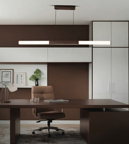 A modern office interior featuring a dark brown wooden desk and matching office chair, with a long rectangular pendant light hanging above. The room has brown and white cabinetry, framed wall art, a small plant, and a gray rug.