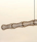 A modern horizontal pendant light with a textured, segmented glass shade suspended by a thin black cord against a plain off-white background.