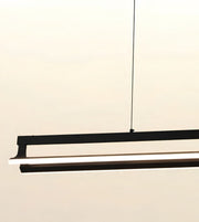 A modern linear LED pendant light fixture in matte black, suspended by a thin black cable against a warm off-white background.