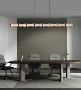 A modern linear pendant light with a textured, segmented wood-look finish hangs over a long conference table with chairs in a minimalist office setting.