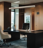A modern office interior with a large black executive desk, black leather office chair, white swivel chair, wood-paneled walls, and a long rectangular ceiling light.