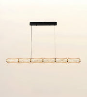 A modern linear pendant light with a textured, translucent bamboo-inspired shade and gold-toned accents, suspended from a slim black ceiling canopy against a warm off-white background.
