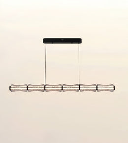 A modern linear chandelier with five textured, translucent glass cylindrical shades suspended from a black rectangular ceiling canopy against a warm off-white background.