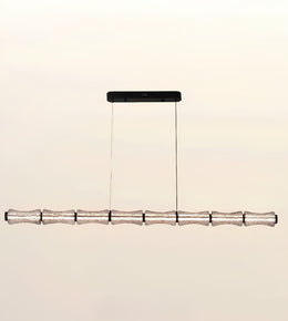 A modern linear chandelier with a textured metallic silver horizontal bar suspended by two thin cables from a black rectangular ceiling mount, shown against a plain light beige background.