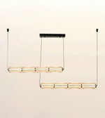 A modern two-light linear chandelier with textured, translucent amber glass shades and gold-toned metal accents, suspended from a slim black ceiling canopy against a warm off-white background.