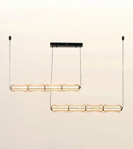 A modern two-light linear chandelier with textured, translucent amber glass shades and gold-toned metal accents, suspended from a slim black ceiling canopy against a warm off-white background.