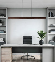 A modern minimalist home office setup featuring a long rectangular wooden pendant light hanging above a white desk with a black office chair. The room is styled symmetrically with light gray shelving units on either side containing books and plants.