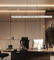 A modern rectangular linear pendant light with a white and gray marble patterned top hangs over a dark wood office desk with a computer monitor, office chair, and other office accessories in a minimalist office setting.