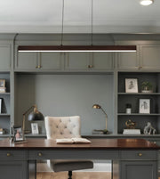 A modern linear pendant light with a dark wood frame hangs over a home office desk setup featuring a tufted cream chair, brass desk lamps, and built-in gray shelving.