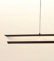 A modern linear LED pendant light with a matte black frame and a warm white glowing diffuser, suspended by a thin black cord against a plain off-white wall.