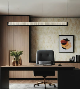 A modern office interior featuring a long rectangular linear pendant light hanging above a desk with a black office chair. The room has light wood paneling, beige patterned wallpaper, framed abstract wall art, and a gray rug.