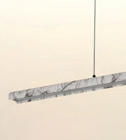 A modern linear pendant light with a white and gray marble-patterned outer shell and a silver metal frame, suspended by a thin black cord against a warm off-white background.