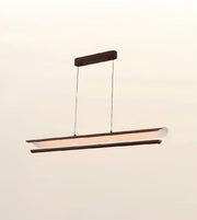 A modern linear LED pendant light with a dark wood frame and a curved, translucent white diffuser, suspended from a matching dark wood ceiling canopy against a plain, light beige wall.
