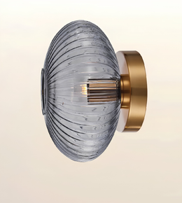 A modern wall-mounted light fixture with a ribbed, oval-shaped clear glass shade and a brushed gold metal wall mount, shown against a soft beige gradient background.
