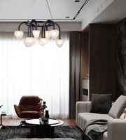 Modern multi-light chandelier with frosted glass globe shades and a black metal frame, hanging in a contemporary living room with a sofa, armchair, and large window.