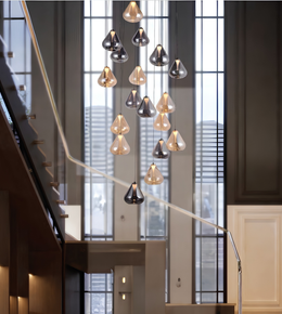 A modern multi-light cluster chandelier with teardrop-shaped glass pendant bulbs in black and smoked glass, hanging in front of a tall window in a contemporary interior staircase setting.
