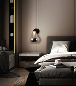 A modern wall-mounted glass pendant light with a black wall mount and a warm glowing bulb, hanging on a taupe wall in a contemporary bedroom setting.